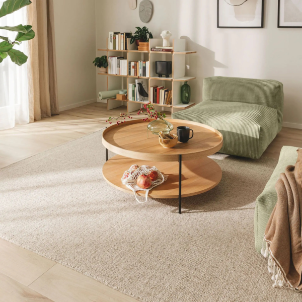 Ein Bild aus dem Taiga Store von taiga.green. Ein modernes Wohnzimmer mit einem runden Couchtisch aus Holz, auf dem eine Teekanne, eine Tasse und eine Schale mit Obst stehen. Es gibt einen grünen Loungesessel, Bücherregale, einen weichen Teppich und Sonnenlicht, das durch transparente Vorhänge fällt. Jetzt bestellen im Eco Shop von www.taiga.green. Hier nachhaltig einkaufen mit dem Taiga Eco Rating