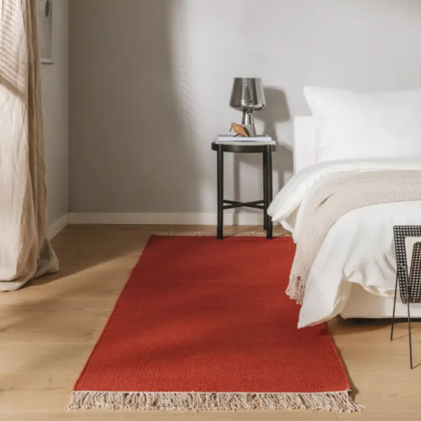 Ein Bild aus dem Taiga Store von taiga.green. Ein gemütliches Schlafzimmer mit einem weißen Bett, einer beigen Decke und einem leuchtend roten Teppich mit Fransen auf hellem Holzfußboden. Ein kleiner schwarzer Beistelltisch mit einer Lampe, einem Buch und einer Brille steht vor einer hellgrauen Wand. Jetzt bestellen im Eco Shop von www.taiga.green. Hier nachhaltig einkaufen mit dem Taiga Eco Rating