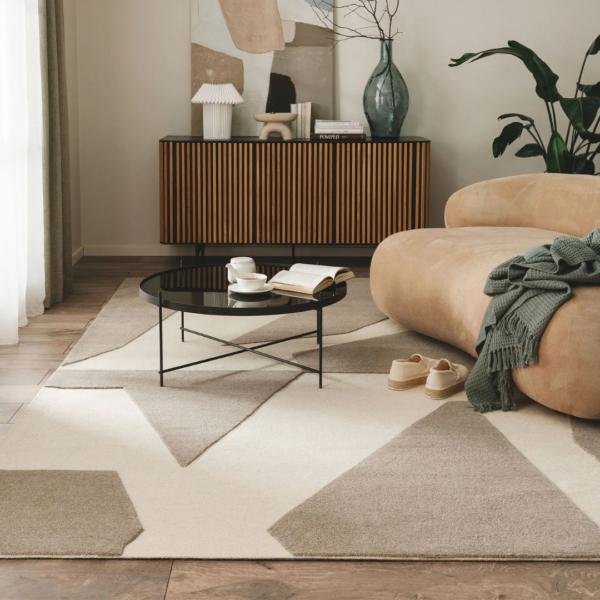 Ein Bild aus dem Taiga Store von taiga.green. Ein modernes Wohnzimmer mit einem beigefarbenen, geschwungenen Sofa, einer grünen Decke, einem runden, schwarzen Couchtisch, einem offenen Buch, einer Tasse, Hausschuhen, einem geometrischen Teppich, einem hölzernen Sideboard, Dekorationsartikeln, abstrakter Wandkunst und einer großen Grünpflanze. Jetzt bestellen im Eco Shop von www.taiga.green. Hier nachhaltig einkaufen mit dem Taiga Eco Rating
