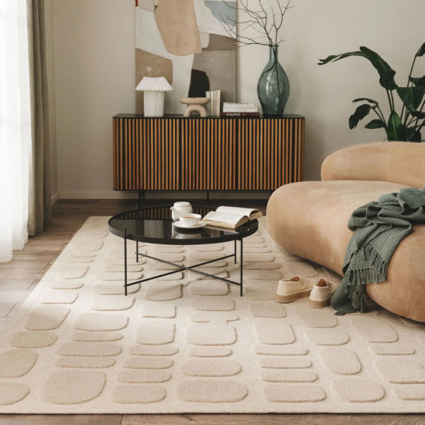 Ein Bild aus dem Taiga Store von taiga.green. Ein modernes Wohnzimmer mit einem beigefarbenen geschwungenen Sofa, einer grünen Decke, gewebten Hausschuhen, einem runden schwarzen Couchtisch, einem aufgeschlagenen Buch, Tassen, einem abstrakten Teppich und einem hölzernen Sideboard mit Vasen, Büchern und Dekoration vor einer hellen Wand. Jetzt bestellen im Eco Shop von www.taiga.green. Hier nachhaltig einkaufen mit dem Taiga Eco Rating