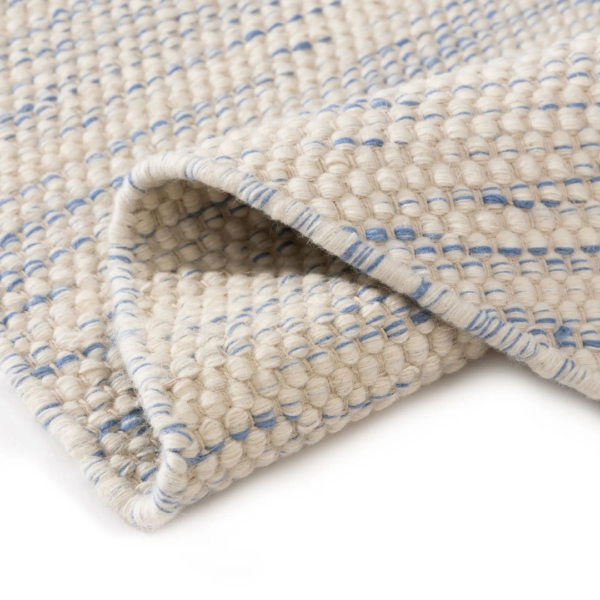 Ein Bild aus dem Taiga Store von taiga.green. Eine Nahaufnahme eines gewebten Teppichs in Creme- und Blautönen, die seine strukturierte Oberfläche und die gefaltete Ecke zeigt. Jetzt bestellen im Eco Shop von www.taiga.green. Hier nachhaltig einkaufen mit dem Taiga Eco Rating