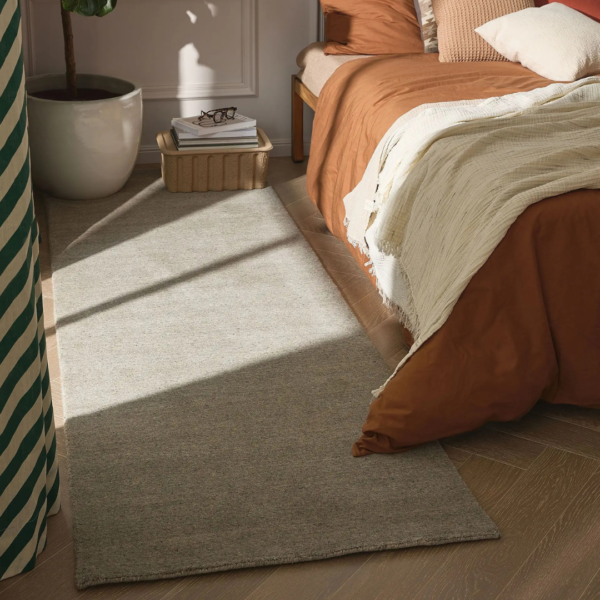 Ein Bild aus dem Taiga Store von taiga.green. Eine gemütliche Schlafzimmerecke mit einem Bett mit rostfarbener Bettwäsche und einer cremefarbenen Decke, einem weichen grauen Teppich auf Holzboden, einem kleinen Nachttisch mit Gläsern und Büchern und einer Topfpflanze im Hintergrund. Jetzt bestellen im Eco Shop von www.taiga.green. Hier nachhaltig einkaufen mit dem Taiga Eco Rating
