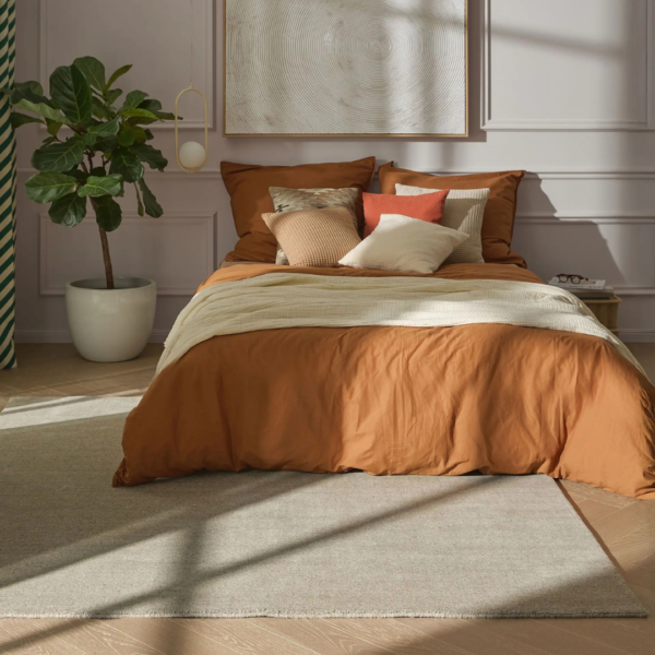 Ein Bild aus dem Taiga Store von taiga.green. Ein ordentlich gemachtes Bett mit rostfarbenem Bettzeug, mehreren Kissen in Beige- und Orangetönen und einer cremefarbenen Decke. Links steht eine Topfpflanze, und Sonnenlicht fällt in das gemütliche, moderne Schlafzimmer mit hellen Wänden und einem großen Teppich. Jetzt bestellen im Eco Shop von www.taiga.green. Hier nachhaltig einkaufen mit dem Taiga Eco Rating
