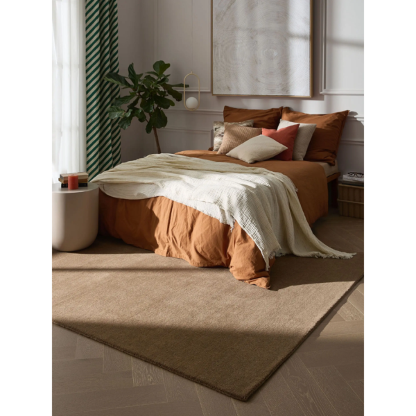 Ein Bild aus dem Taiga Store von taiga.green. Ein gemütliches Schlafzimmer mit einem Bett mit rostfarbener Bettwäsche, weißer Überwurfdecke und mehreren Kissen. Eine grüne Pflanze steht neben dem Bett, und sanftes Sonnenlicht fällt durch ein Fenster mit grünen Vorhängen. Ein beiger Teppich bedeckt den Holzboden. Jetzt bestellen im Eco Shop von www.taiga.green. Hier nachhaltig einkaufen mit dem Taiga Eco Rating
