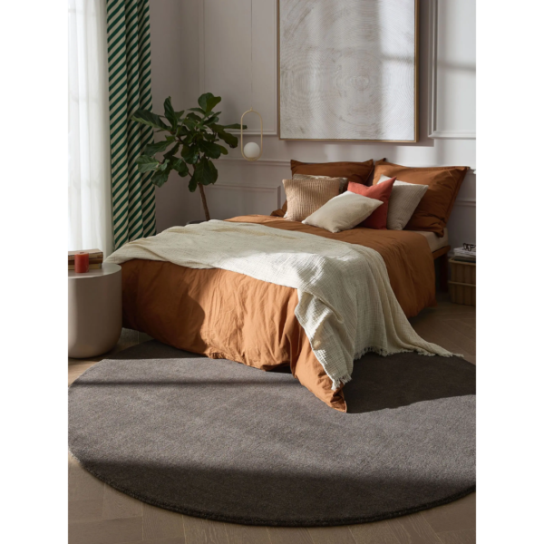 Ein Bild aus dem Taiga Store von taiga.green. Ein gemütliches Schlafzimmer mit einem Bett, das mit braunen Laken, einer cremefarbenen Decke und verschiedenen Kissen bezogen ist. Neben dem Bett liegen ein großer grauer runder Teppich und eine Topfpflanze. Das natürliche Licht fällt durch einen grün-weißen Vorhang. Jetzt bestellen im Eco Shop von www.taiga.green. Hier nachhaltig einkaufen mit dem Taiga Eco Rating